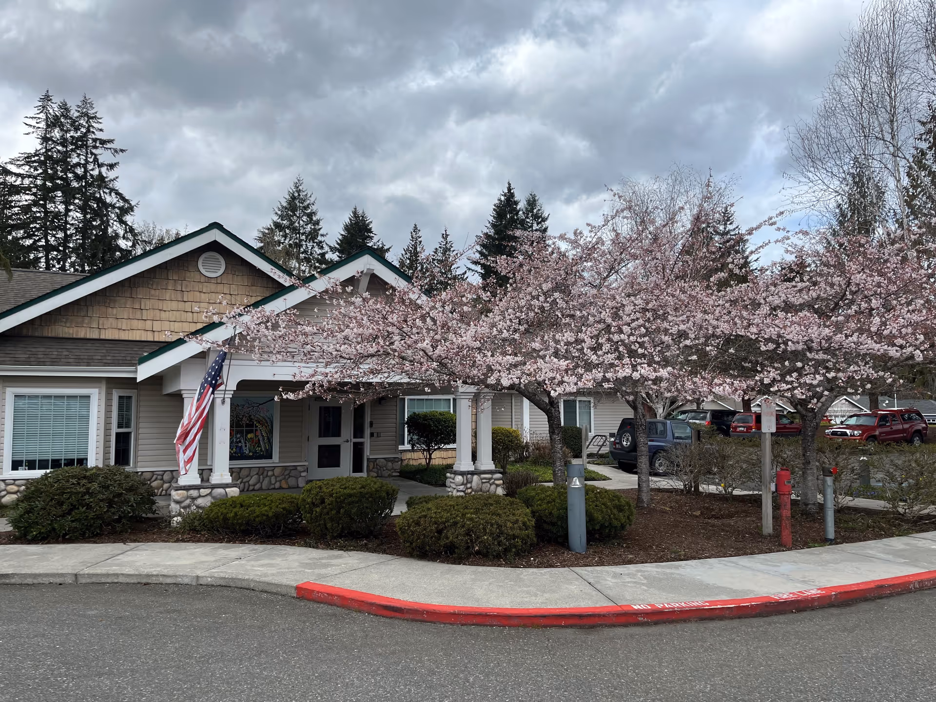Front entrance of a single-story care building with blooming pink cherry trees, an American flag, and parked cars nearby.