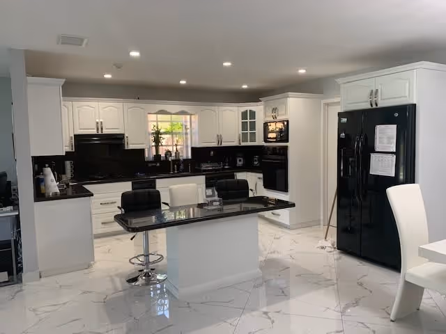 Modern kitchen with white cabinetry, black countertops, and a black refrigerator. There is a central island with a glass top and three chairs, two black and one white. The floor is white with a marble pattern, and there is a window above the sink letting in natural light.