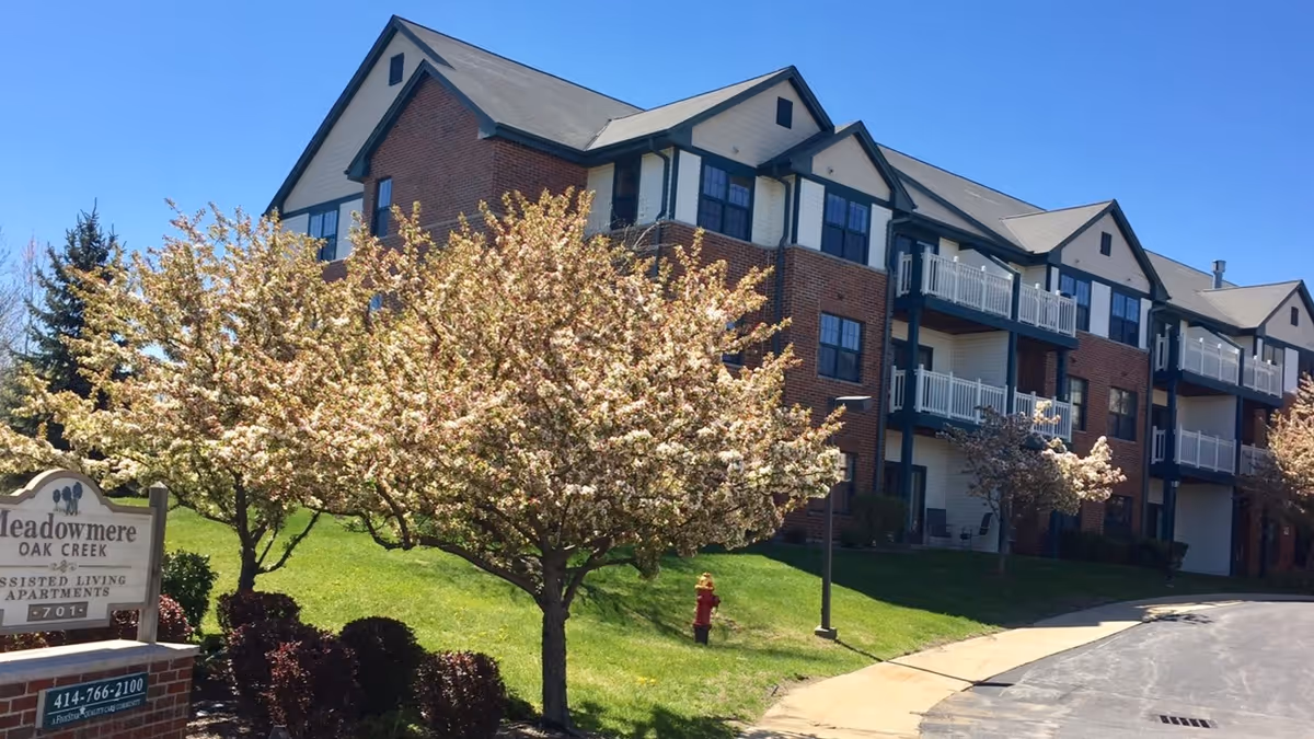 Front exterior of a brick senior living apartment building with balconies, blooming trees and a Meadowmere Oak Creek sign.