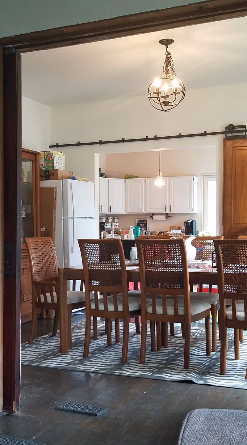 View of a dining area with a wooden table and six chairs on a patterned rug. In the background, there is a kitchen with white cabinets, a white refrigerator, and various kitchen items on the countertops. Two pendant lights hang from the ceiling.
