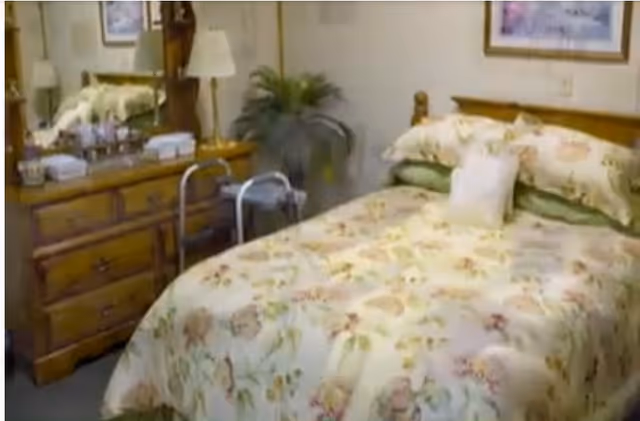 Cozy single bedroom with a floral bedspread, wooden headboard and dresser with mirror, lamp, potted plant, and a walker beside the bed.