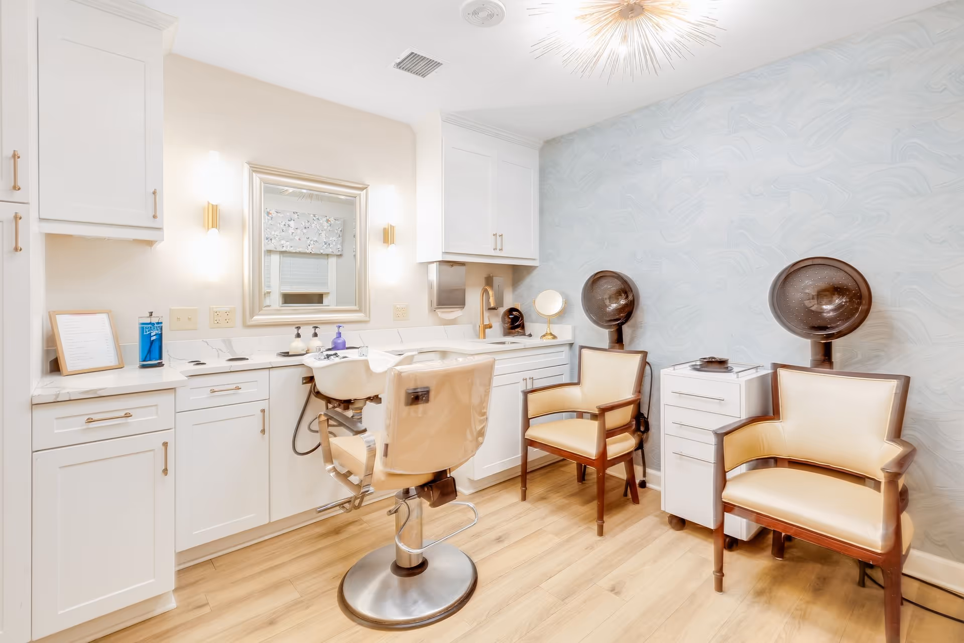 A bright and clean salon room with a hair washing station, a salon chair, two beige armchairs, two hair dryers, white cabinetry, a large mirror, and soft lighting fixtures on the wall.