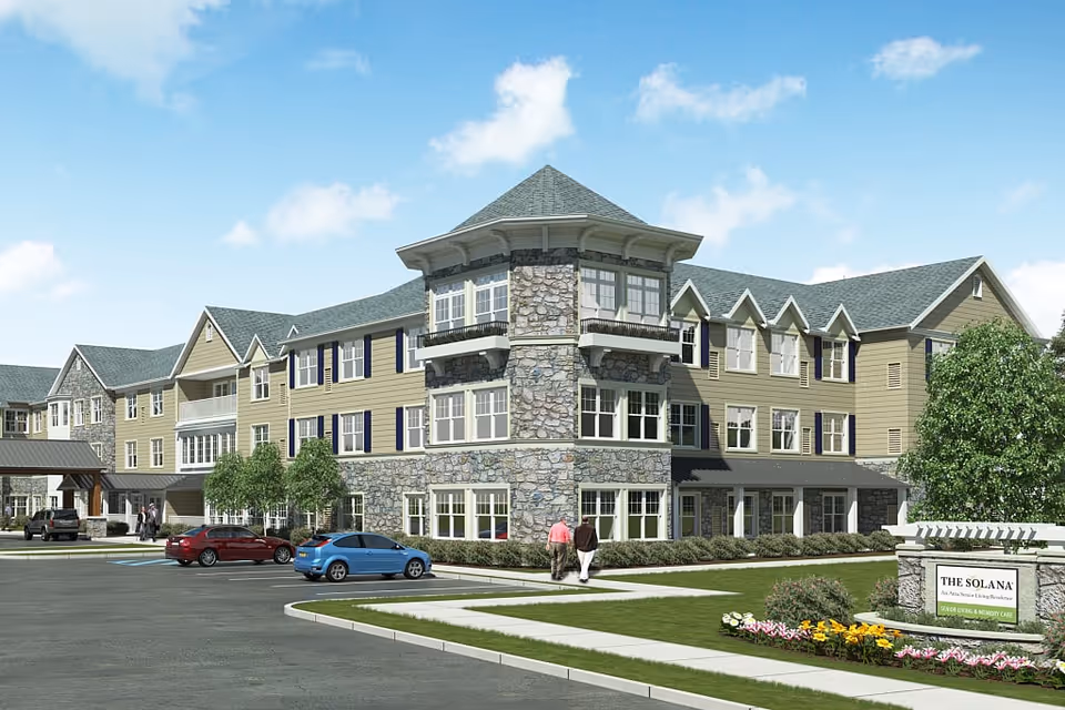 Exterior view of The Solana Doylestown senior living facility showing a large three-story building with stone and beige siding, multiple windows, a parking lot with cars, landscaped greenery, and two people walking on a sidewalk near the entrance.