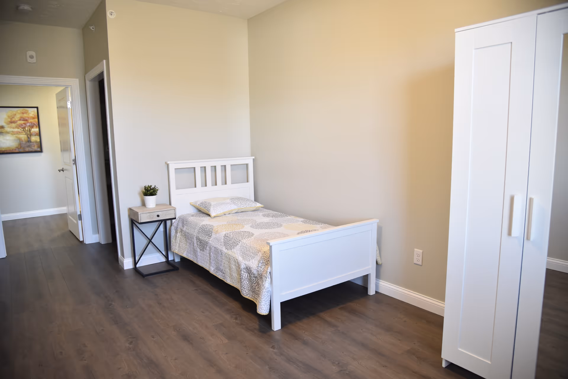 Simple furnished bedroom with a white single bed, nightstand with a small plant, and a white wardrobe on wood floors.