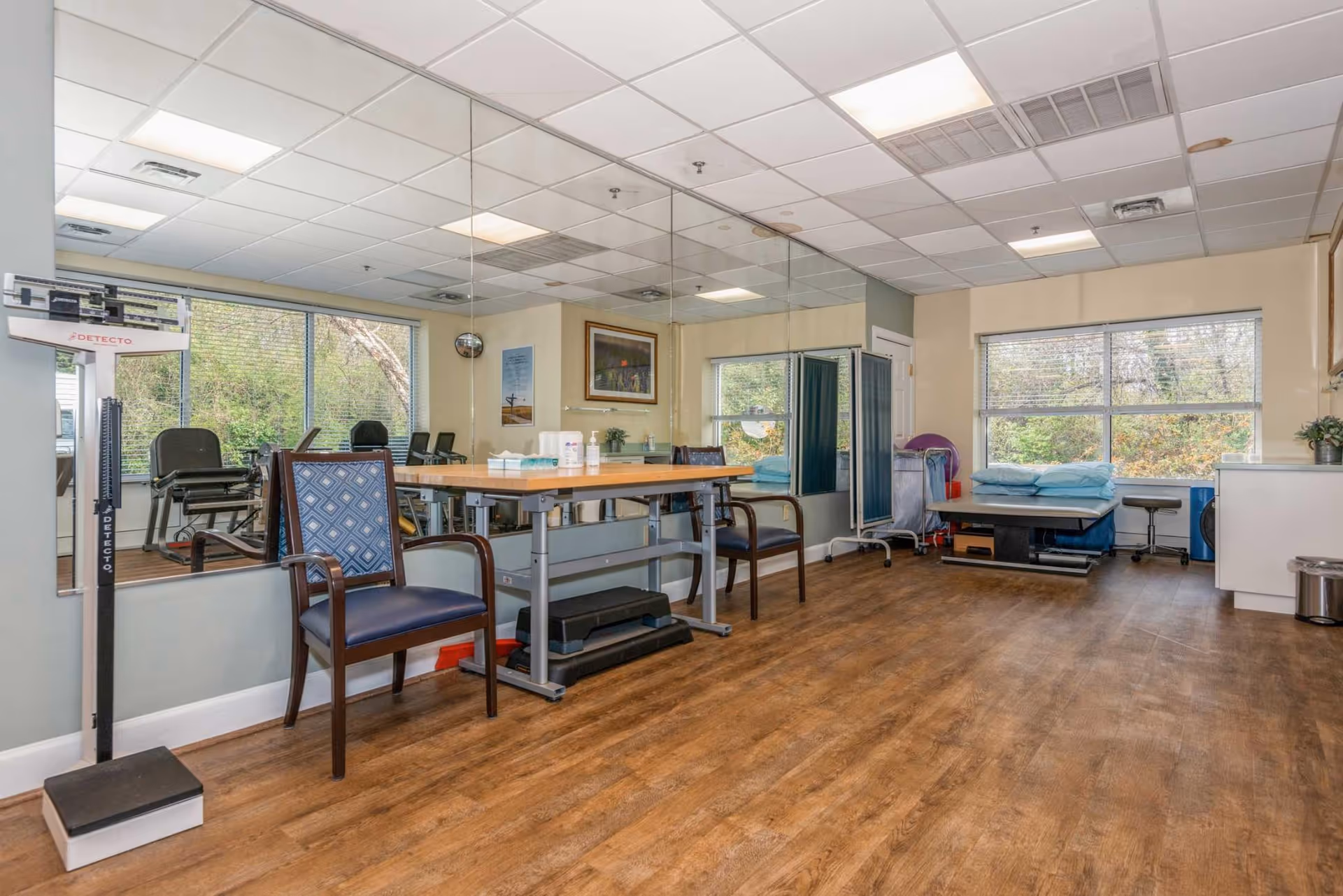 A well-lit therapy or medical examination room with wooden flooring, a large wall mirror, two chairs, a table with medical supplies, a weight scale, an examination table with pillows, a privacy screen, and large windows showing greenery outside.