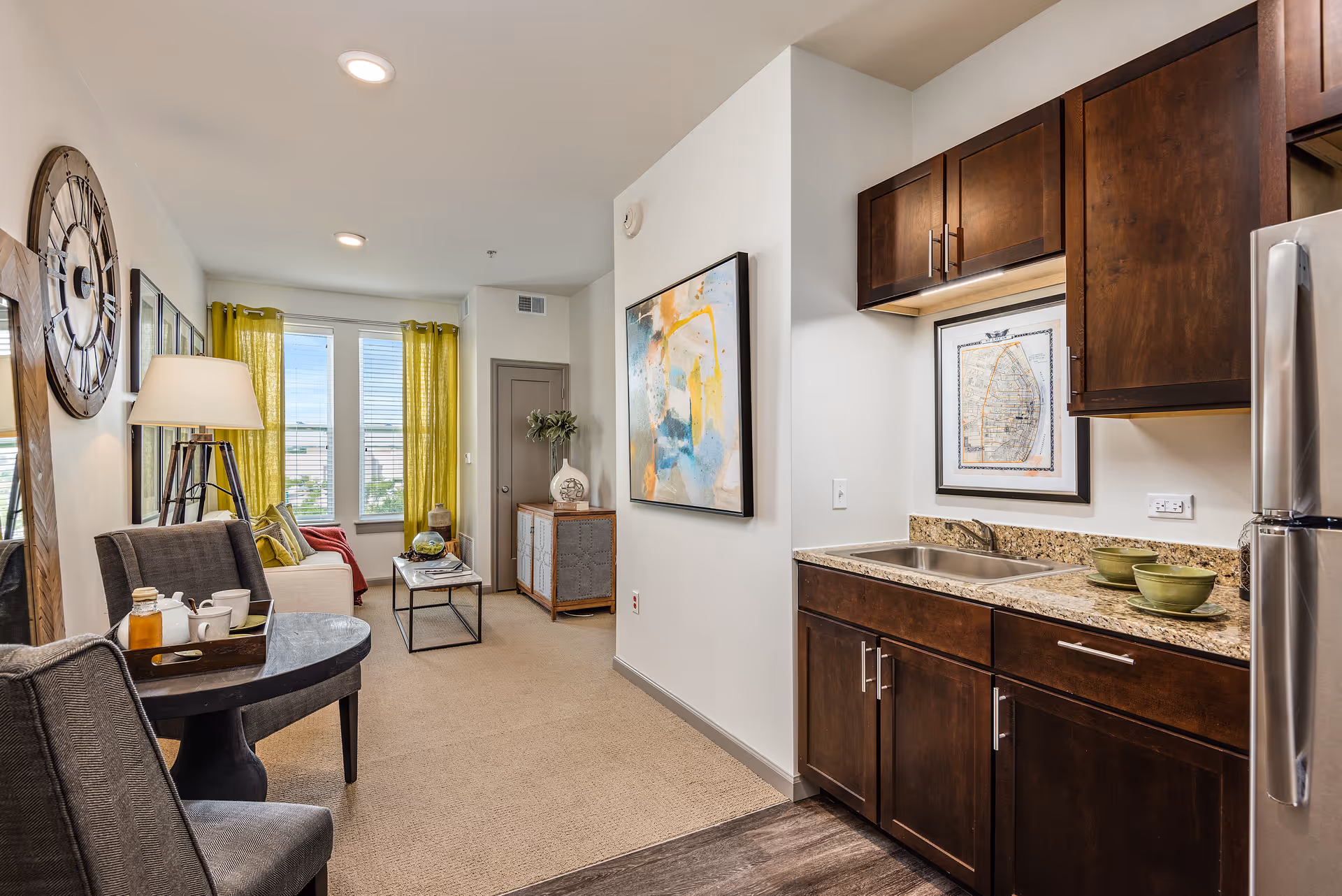 Interior view of a senior living facility apartment showing a small kitchen area with dark wood cabinets, a granite countertop, a sink, and a stainless steel refrigerator. Adjacent to the kitchen is a living area with a beige carpet, a white sofa, a coffee table, a floor lamp, and bright yellow curtains on the window. The walls are decorated with a large clock, framed pictures, and a colorful abstract painting.