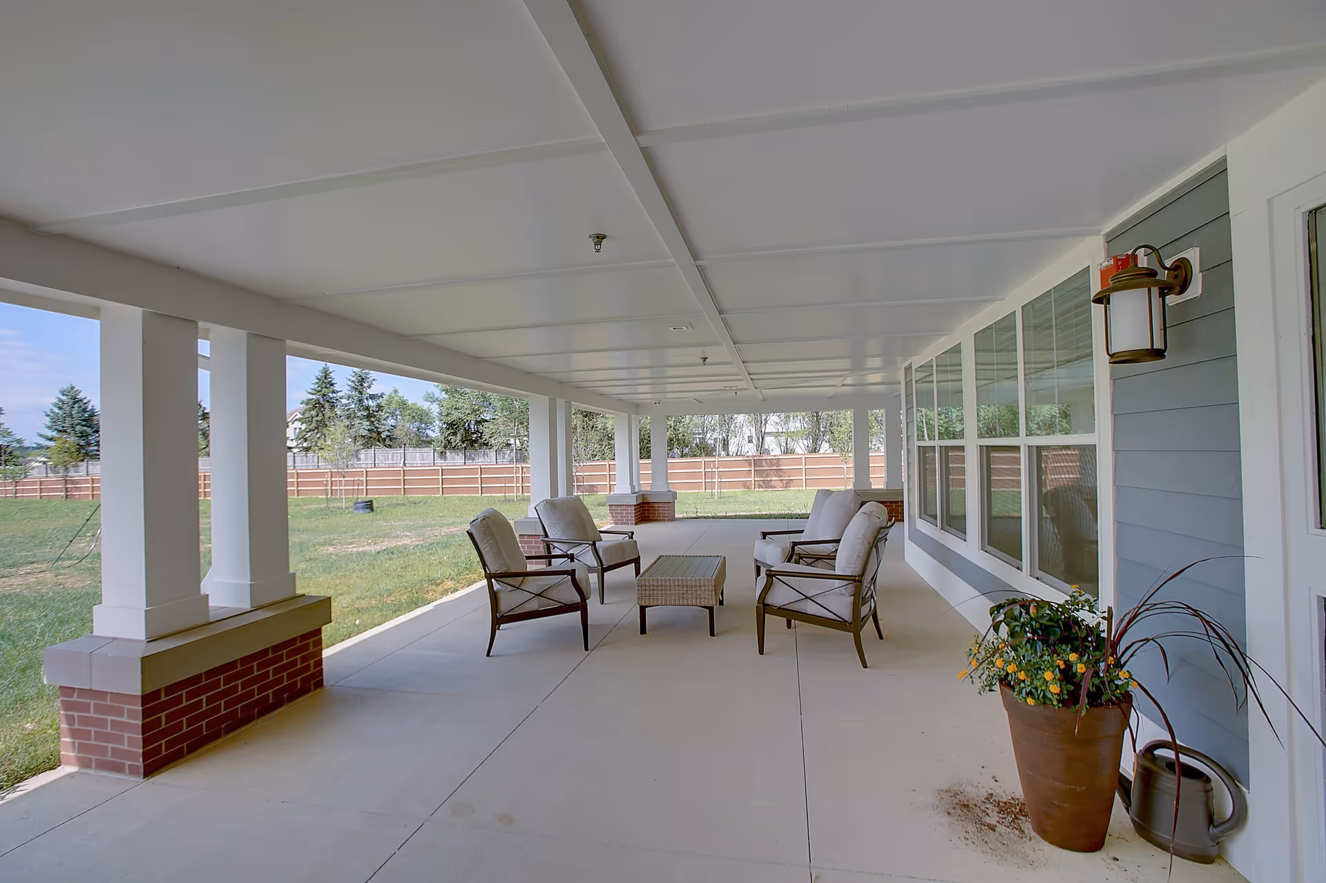 Covered outdoor patio area with four cushioned chairs arranged around a rectangular wicker coffee table. The patio has a white ceiling with beams and brick pillars supporting it. There is a large potted plant and a watering can near the blue siding wall with multiple windows. The patio overlooks a grassy fenced yard with trees in the background.