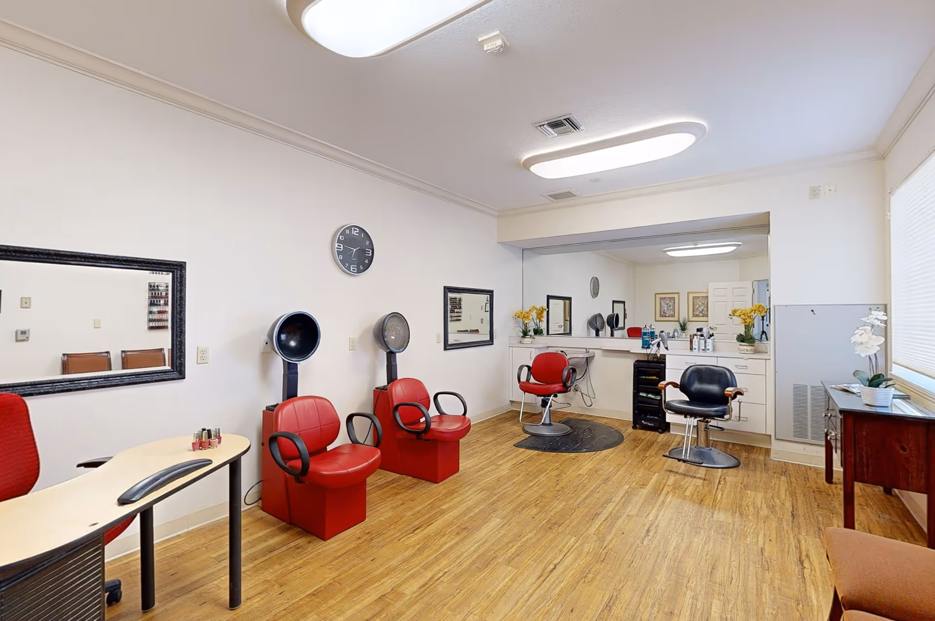 Interior view of a salon area in a senior living facility featuring red salon chairs, hair dryers, mirrors on the walls, a wooden floor, and a reception desk with a red chair. The room is well-lit with ceiling lights and has decorative flowers on the counter and a side table.