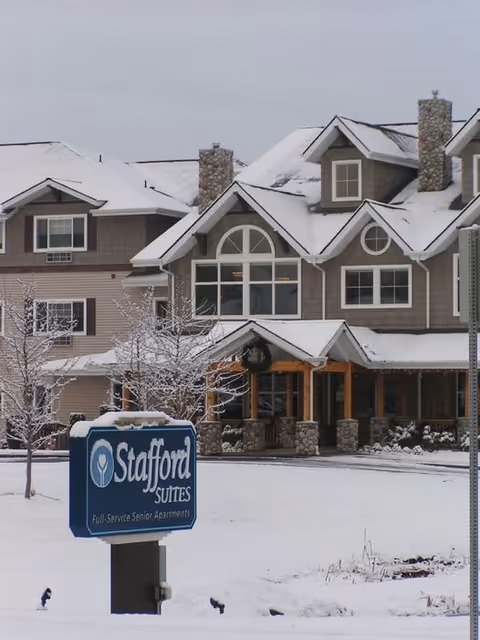 Exterior view of a senior living facility building in winter with snow covering the ground and rooftops. The building has multiple windows, stone chimneys, and a covered entrance with wooden pillars. A blue sign in the foreground reads 'Stafford Suites Full-Service Senior Apartments.'