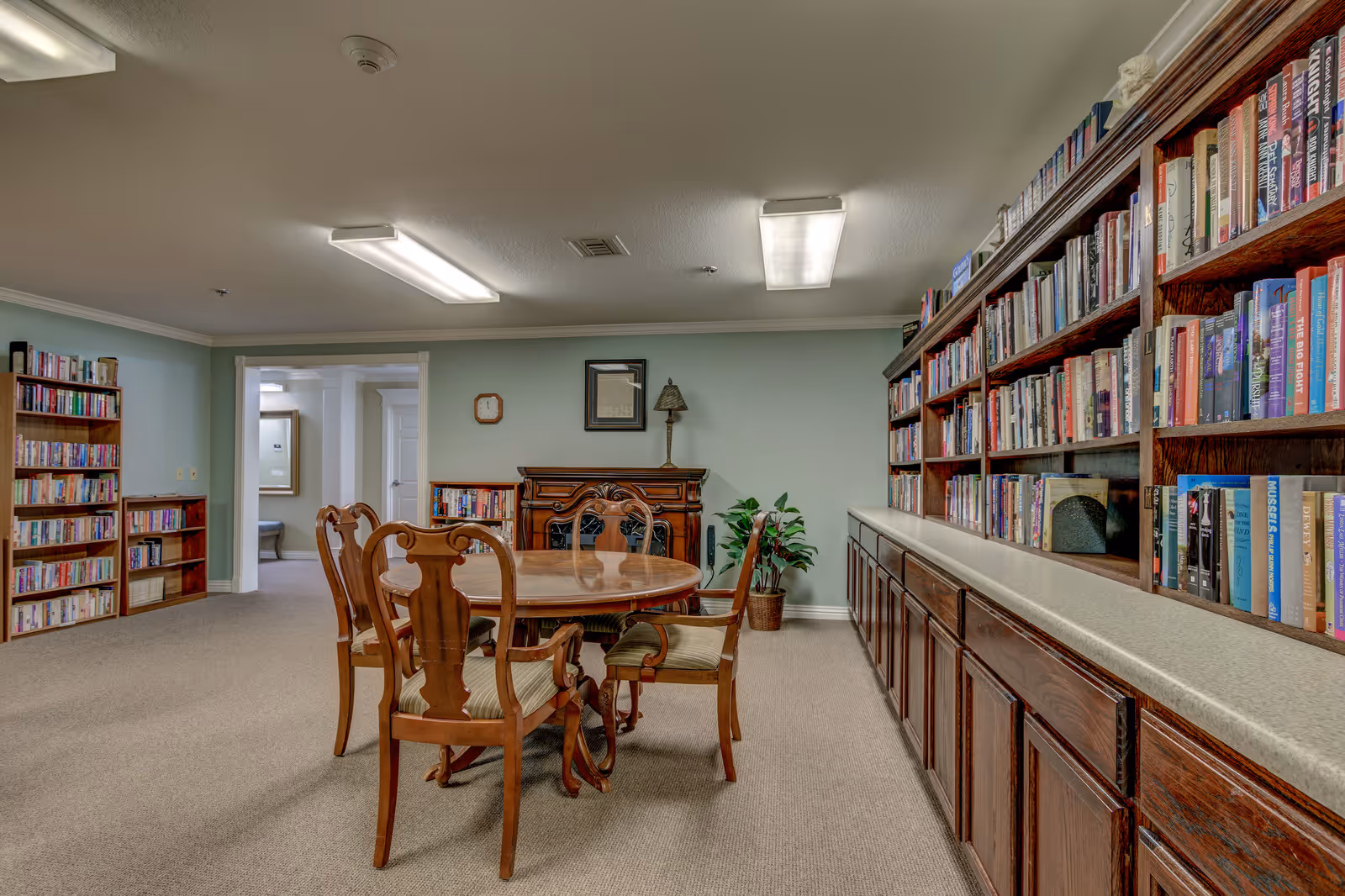 A cozy library room in a senior living community with wooden bookshelves filled with books along the right wall and a smaller bookshelf on the left. In the center, there is a round wooden table surrounded by four wooden chairs with cushioned seats. The room has beige carpet, light green walls, and ceiling lights. A framed picture, a clock, and a lamp are on the far wall above a wooden cabinet, with a potted plant next to it.