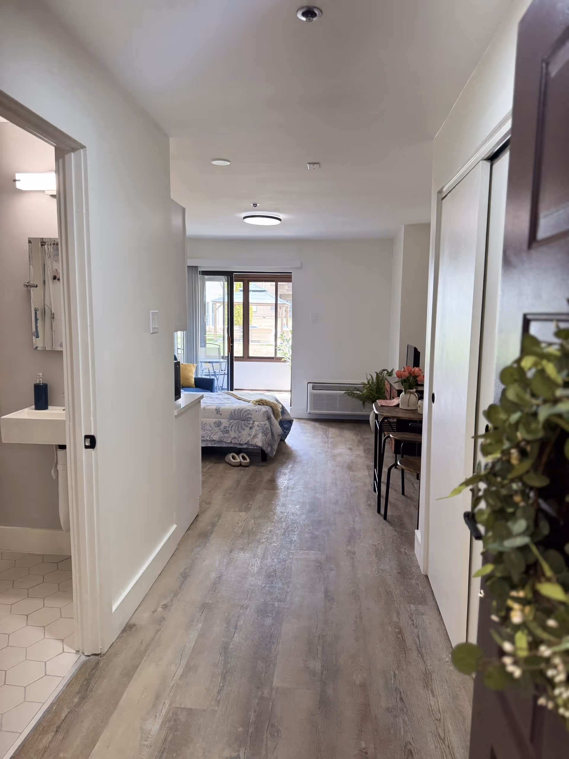 View down a hallway in a senior living facility showing a bathroom on the left with a sink and mirror, a bedroom area with a bed, slippers, and a window at the far end, and a small table with chairs and flowers on the right side. The floor is wood-style laminate and the walls are painted white.