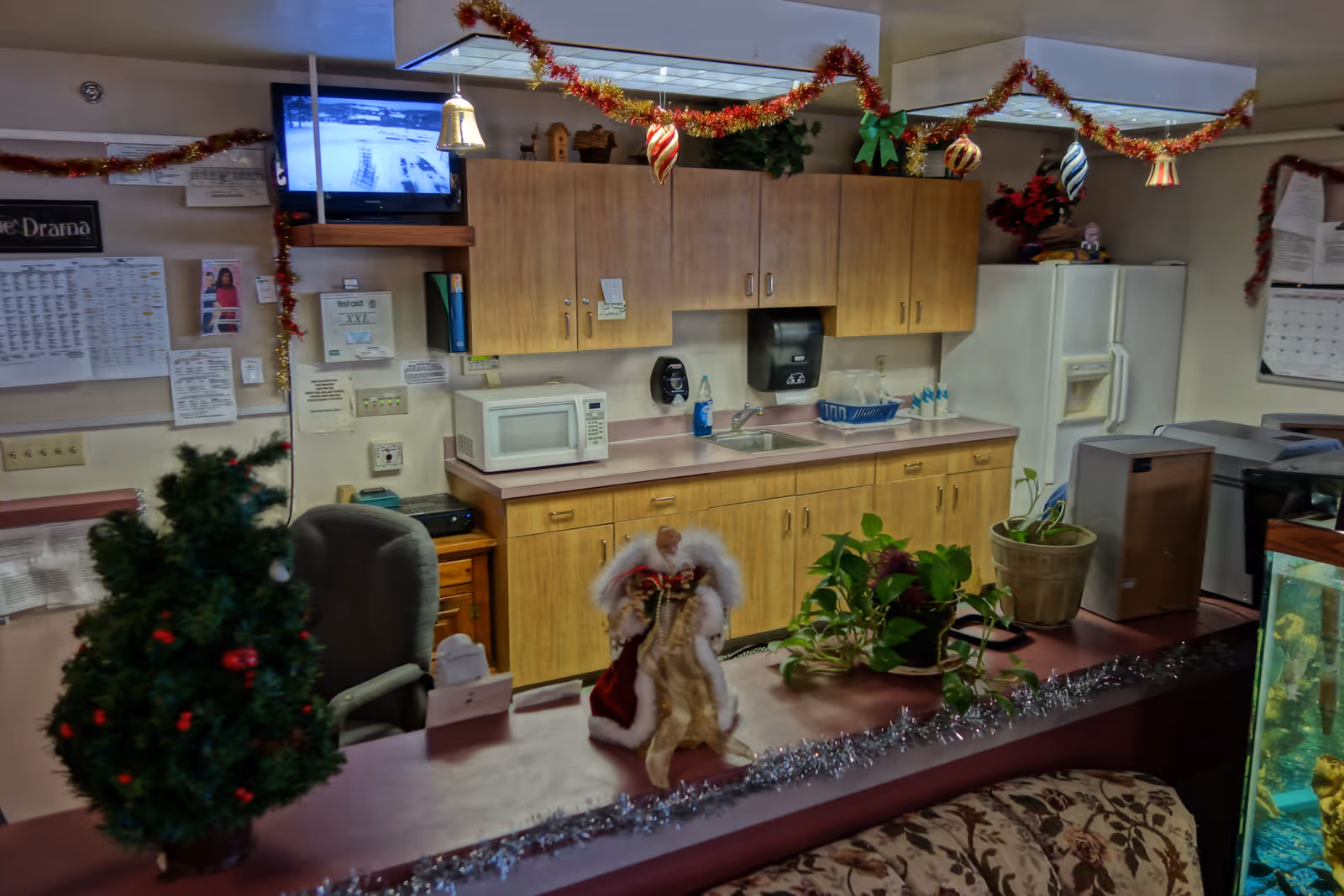 Interior view of a small kitchen area decorated for Christmas with garlands and ornaments hanging from the ceiling. The kitchen has wooden cabinets, a microwave, a sink, a refrigerator, and various plants on the counter. A small decorated Christmas tree and a Santa Claus figurine are on the counter in the foreground. A TV is mounted on the wall showing a black and white image, and various papers and calendars are pinned on the wall.
