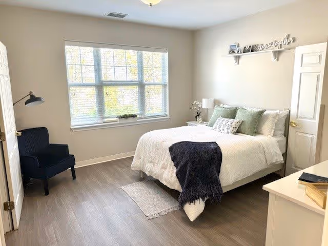 A bright bedroom with a large window letting in natural light. The room features a neatly made bed with white bedding, green and patterned pillows, and a dark throw blanket. There is a black armchair in the corner, a white nightstand with a lamp and decorative items, and a white dresser with books on top. The walls are light-colored, and a shelf above the bed holds framed photos and a decorative 'Together' sign.