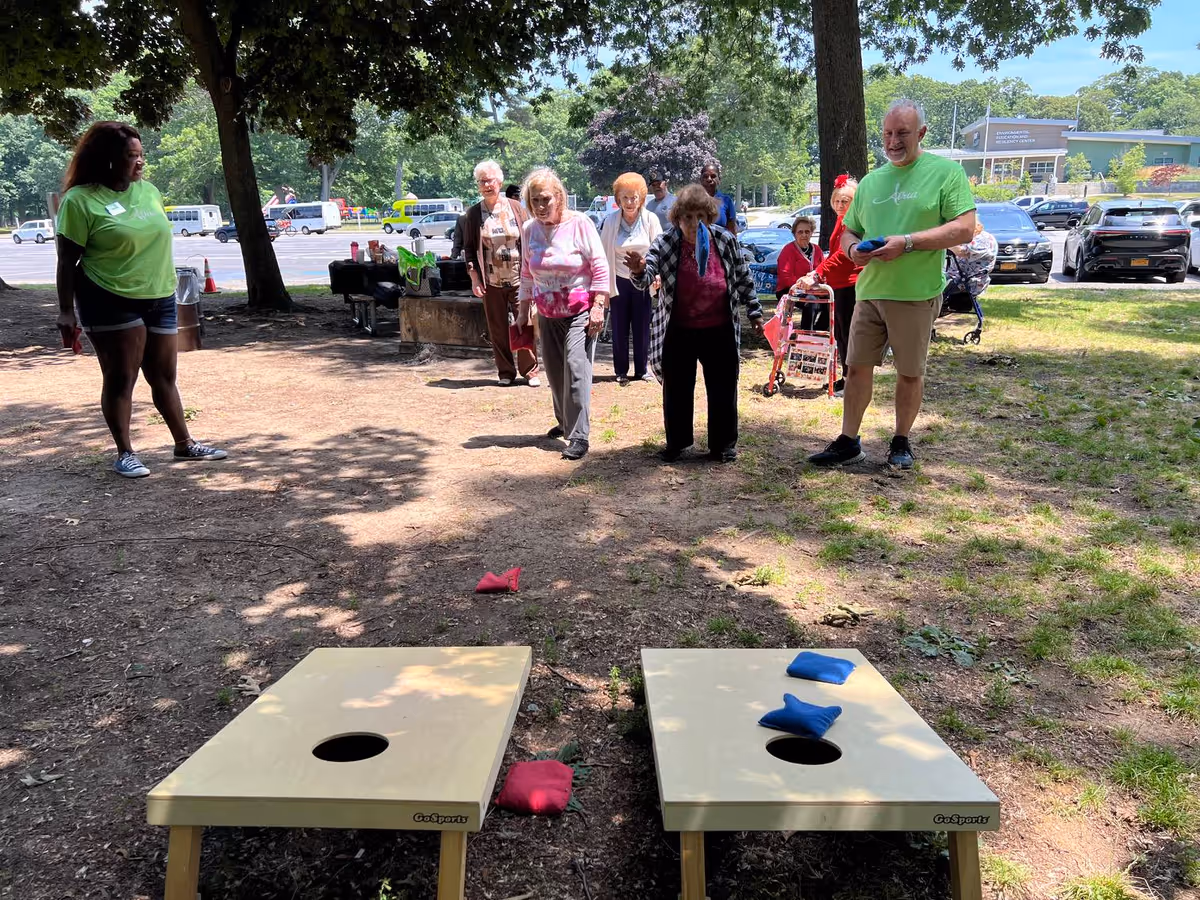 A group of elderly people and two staff members playing cornhole outdoors on a sunny day. Two cornhole boards with bean bags are set up on the ground, and the participants are standing behind the boards, actively engaged in the game. Trees and parked cars are visible in the background.