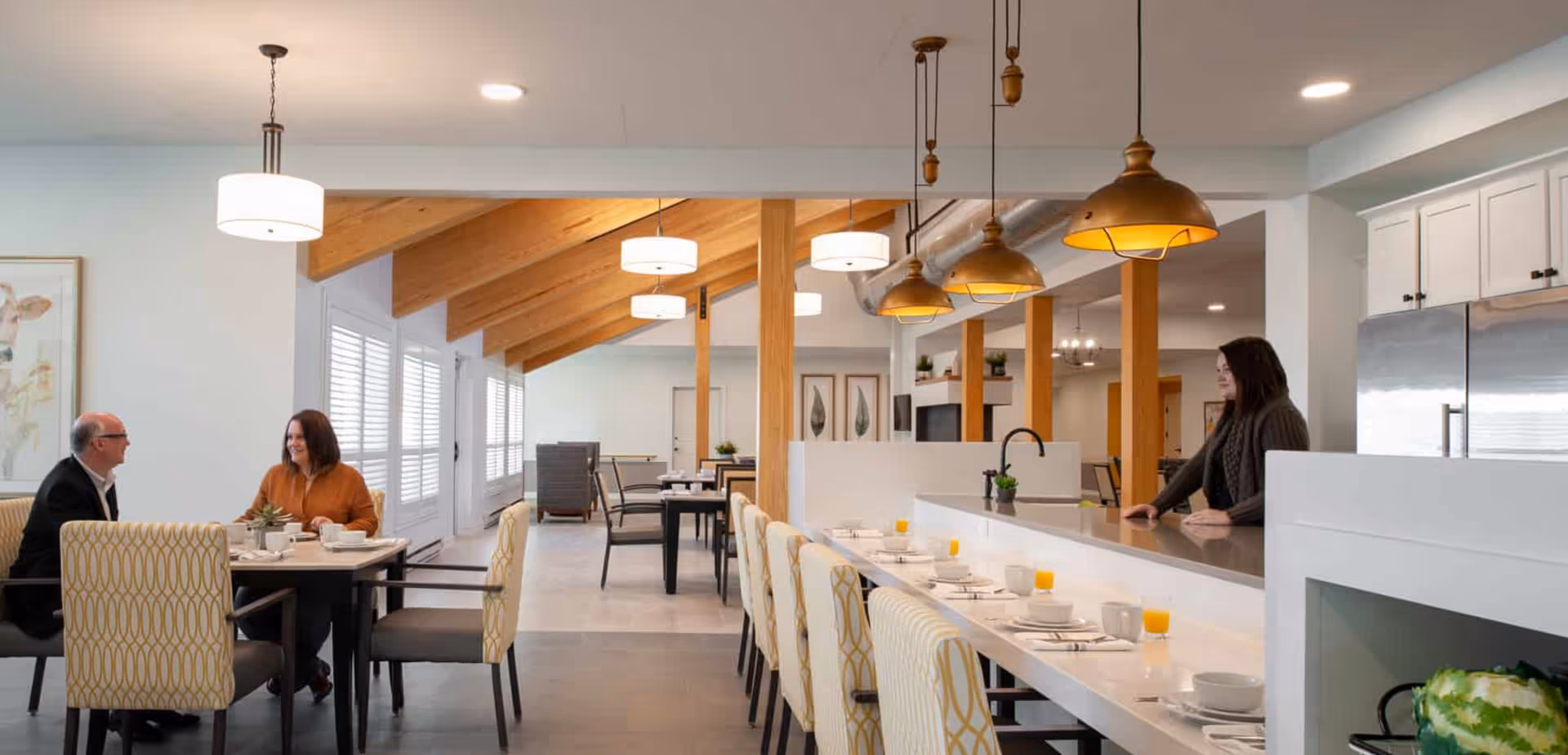 A modern dining area in a senior living facility featuring a long counter with high chairs set with plates, cups, and glasses of orange juice. Two people are seated at a table having a conversation, and a woman stands behind the counter. The room has wooden beams on the ceiling, pendant lights, and large windows with white shutters.
