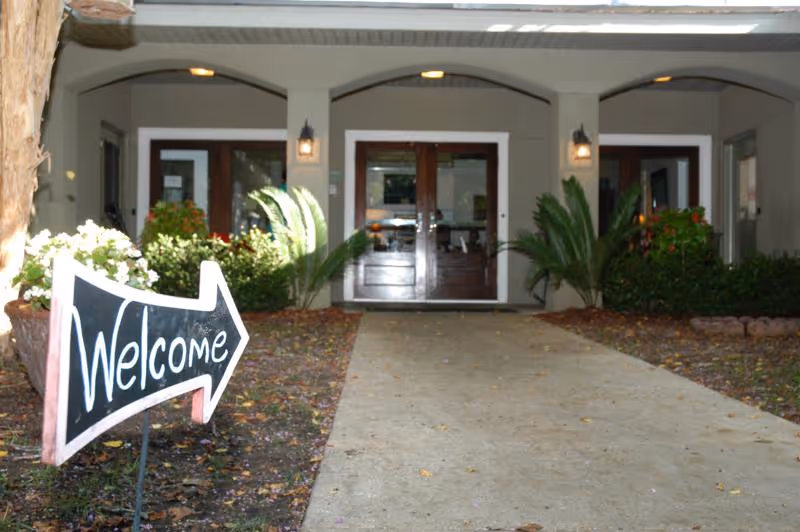 Covered front entrance with a 'Welcome' arrow sign pointing down a walkway to double wooden doors flanked by plants.