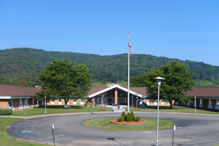 Front exterior view of Sena Kean Nursing and Rehabilitation Center, a single-story brick building with a central entrance, surrounded by trees and hills under a clear blue sky. An American flag flies on a flagpole in front of the building, with a circular driveway and landscaped greenery.