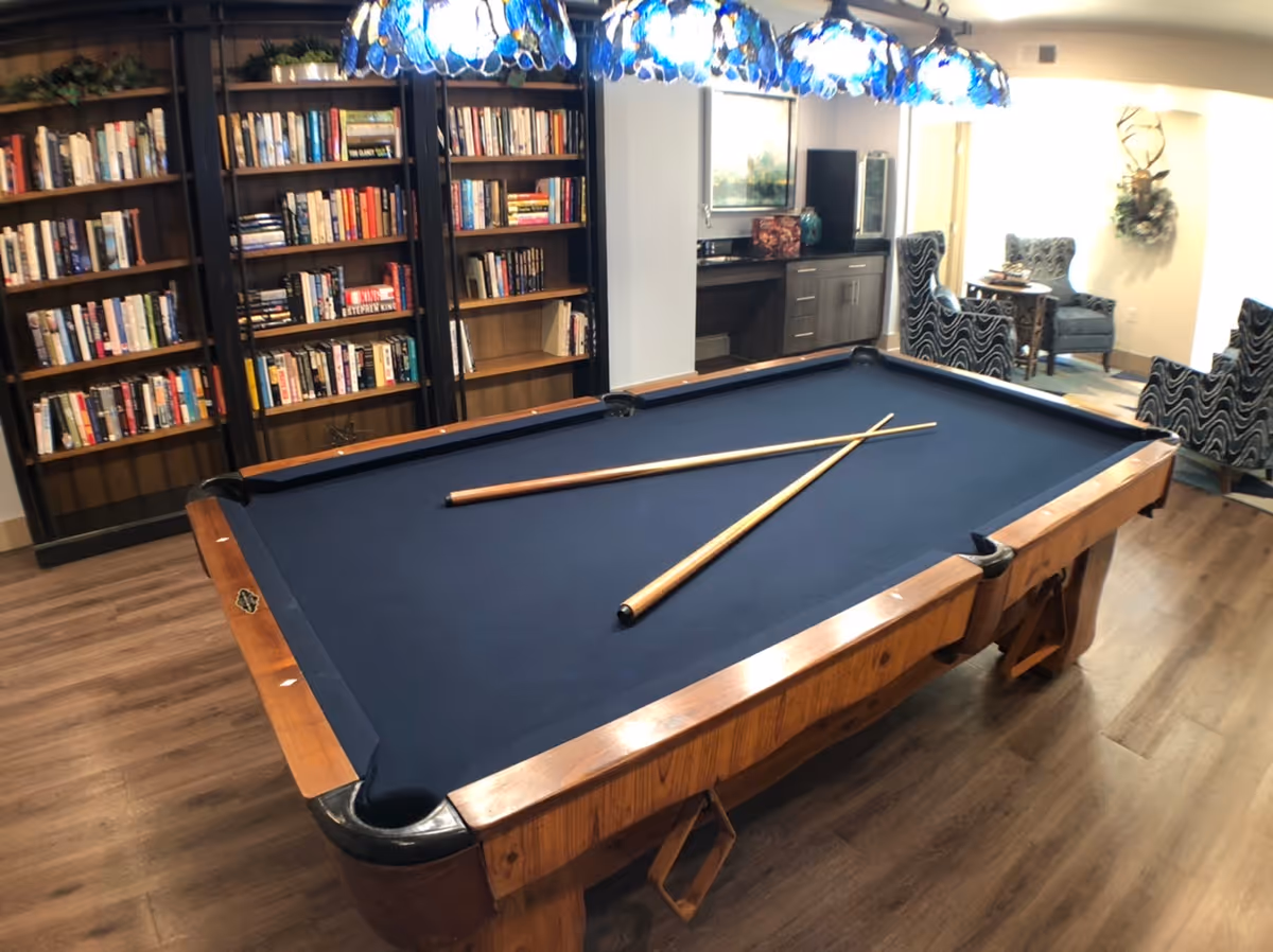 A pool table with navy blue felt and two cues in a lounge area with bookshelves and seating.