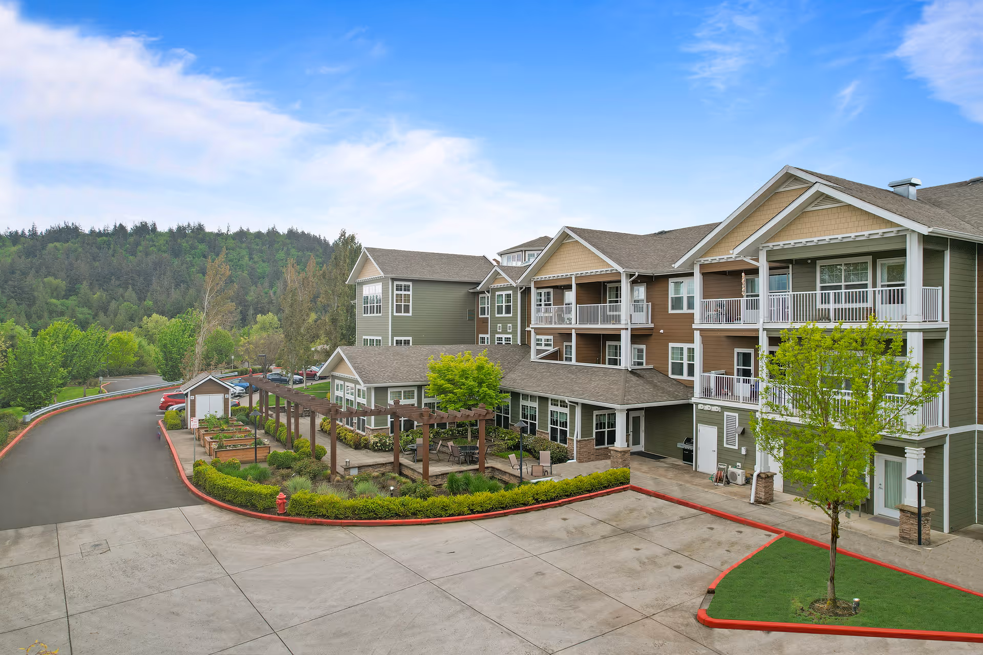 Exterior view of a multi-story senior living building with balconies, a landscaped courtyard and parking area under a blue sky.