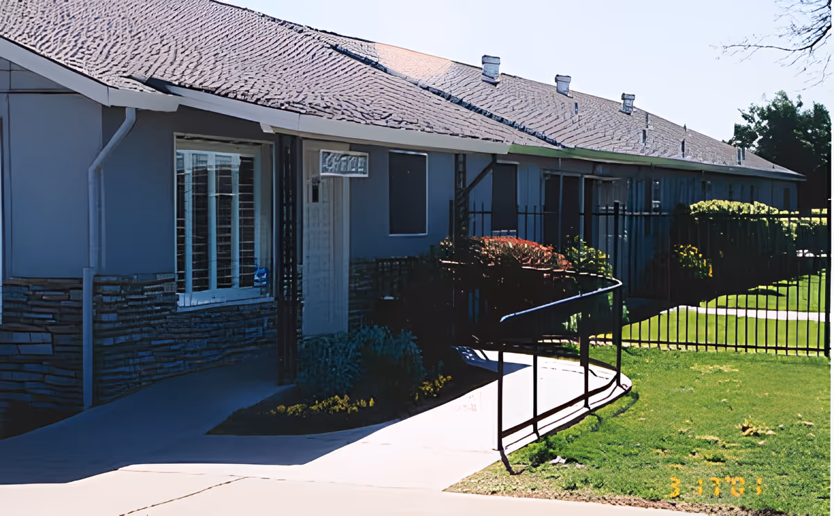 Exterior view of a single-story building with a sloped roof, multiple windows, and a white door. There is a black metal fence and a concrete ramp leading to the entrance. The lawn is green and well-maintained with some bushes near the building.