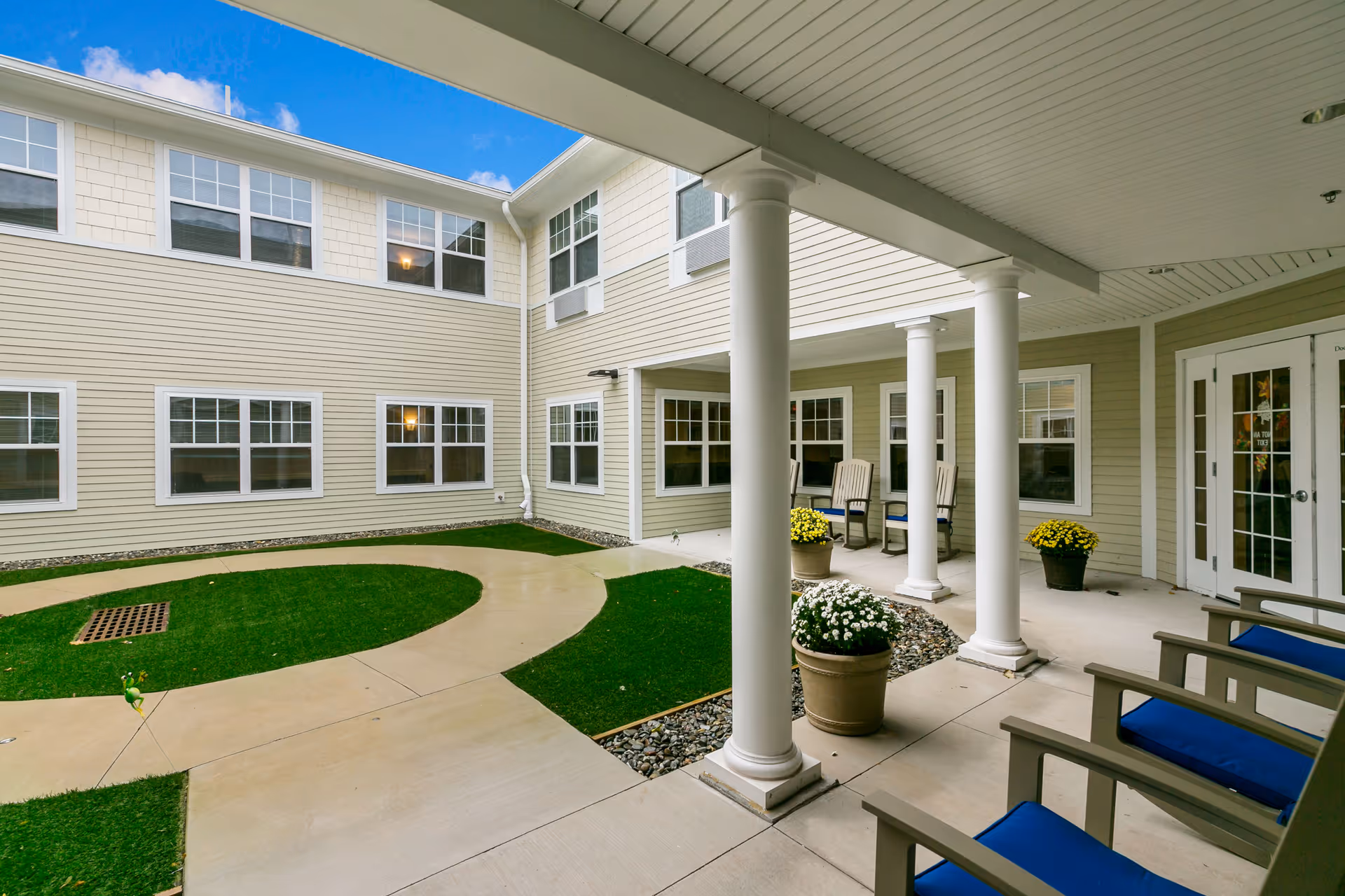 Outdoor courtyard area of a senior living facility with a covered patio supported by white columns, beige siding walls, multiple windows, potted flowers, and several chairs with blue cushions arranged along the patio.