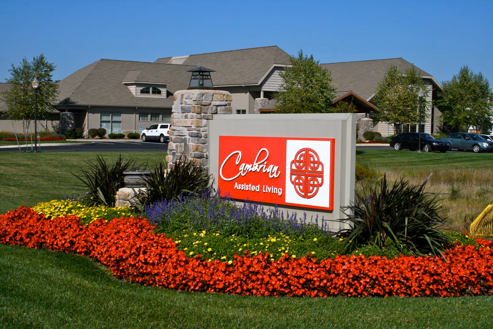 Exterior view of Cambrian Assisted Living and Memory Care Tecumseh building with a large sign in front surrounded by colorful flower beds and green grass under a clear blue sky.