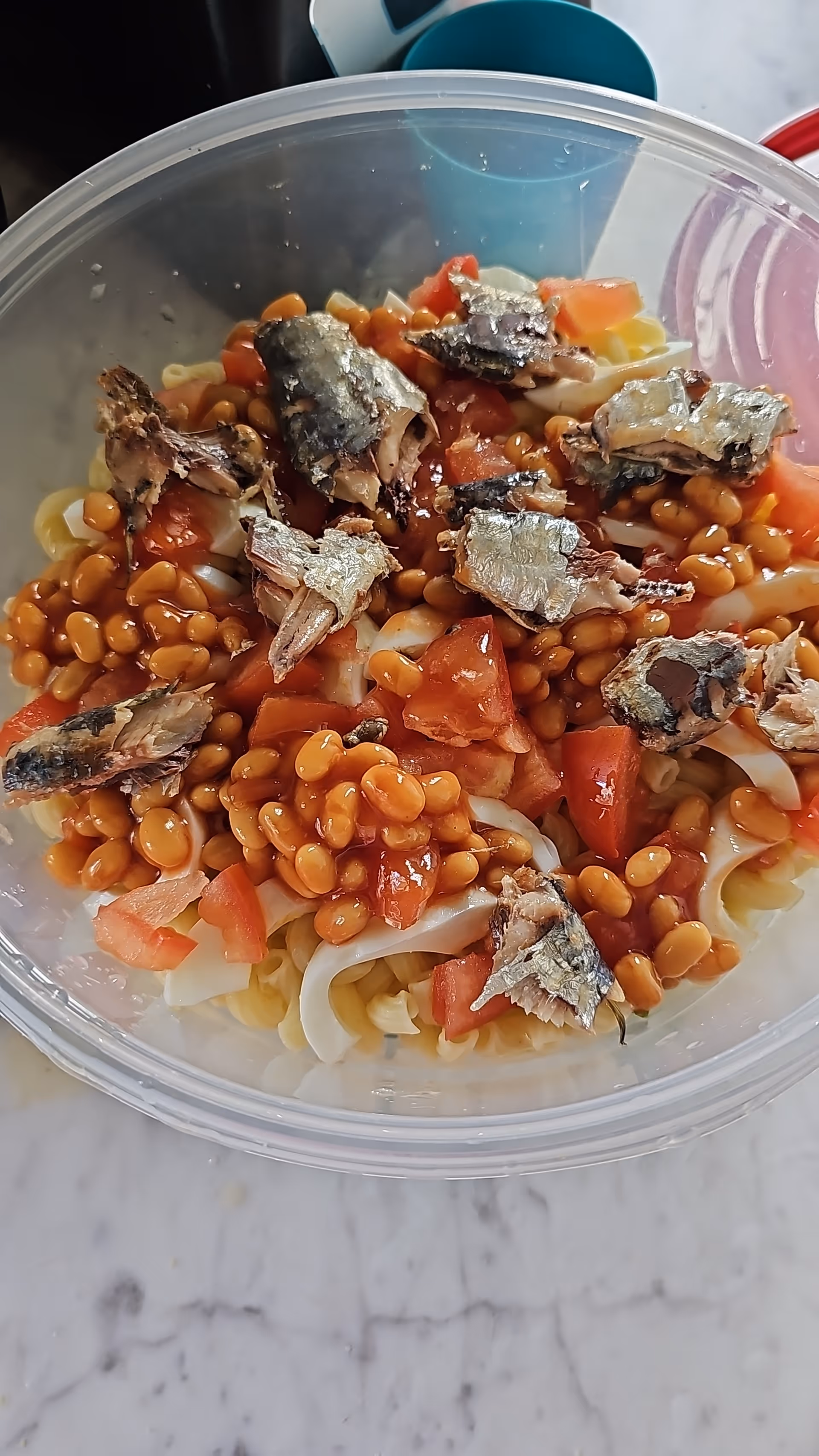 A clear plastic bowl containing a mixture of cooked pasta, baked beans, chopped tomatoes, and pieces of sardines on a marble surface.