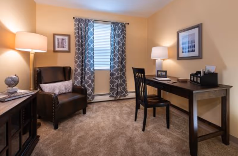 A cozy room with beige walls and carpeted floor featuring a dark wooden desk with a chair, a table lamp, and office supplies. To the left, there is a dark leather armchair with a cushion, a floor lamp, and a small cabinet with decorative items. A window with patterned curtains is centered on the back wall.
