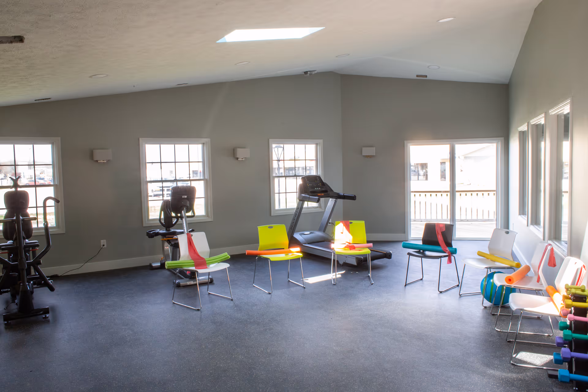 A bright exercise room with several chairs arranged in a semi-circle, each chair equipped with colorful resistance bands and foam rollers. The room has large windows letting in natural light, a treadmill, stationary bike, and a rack of dumbbells in the corner. The walls are painted light gray and the floor is covered with dark rubber gym flooring.