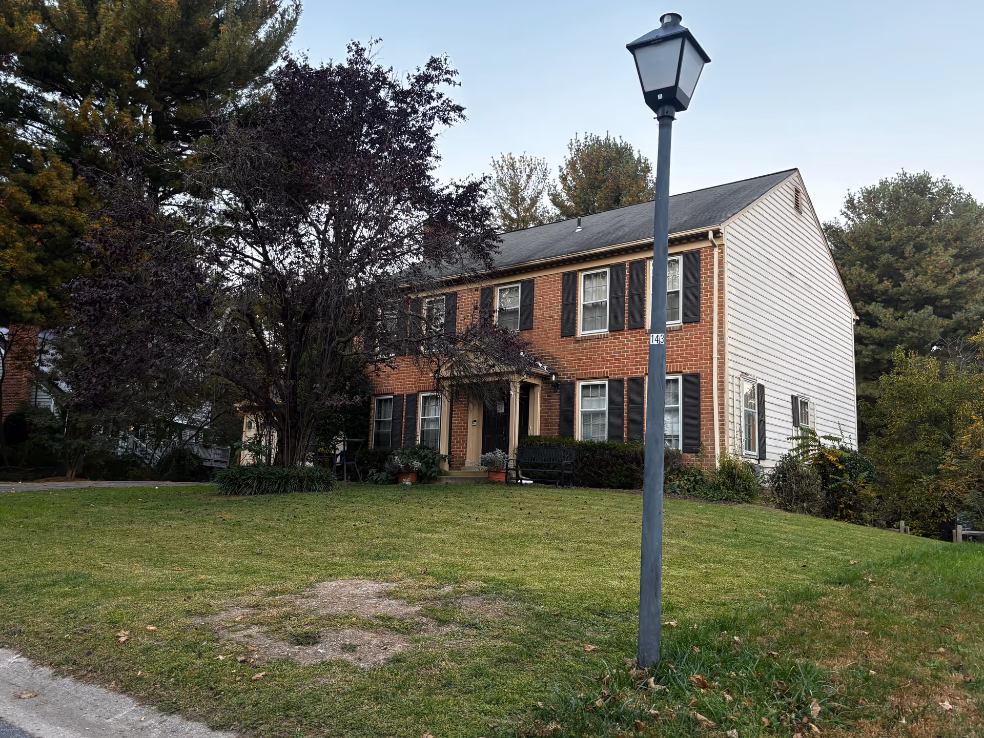 A two-story brick and siding house with black shutters, surrounded by trees and a grassy lawn. A black lamp post stands in the foreground near the street.