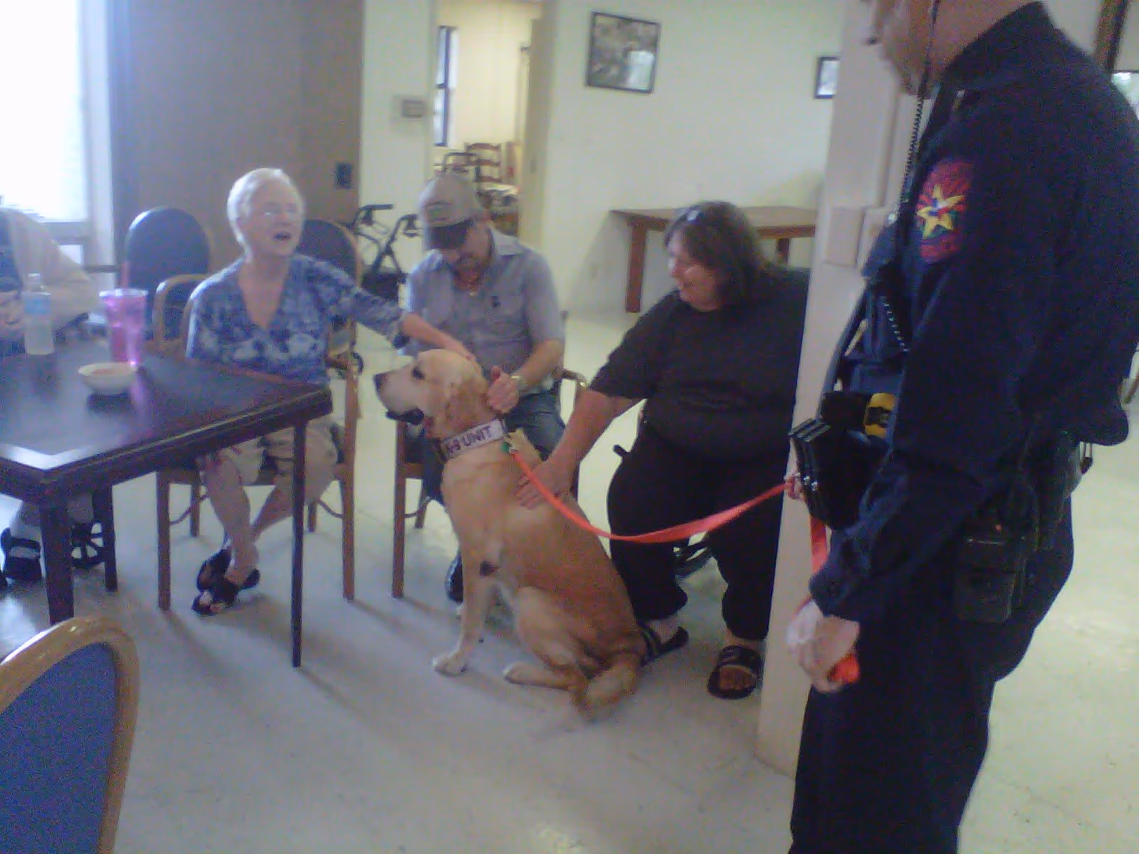 Several elderly people sitting around a table in a room, petting a large golden retriever service dog wearing a vest labeled 'SECURITY'. A uniformed officer is holding the dog's leash. The setting appears to be a communal area in an assisted living facility.