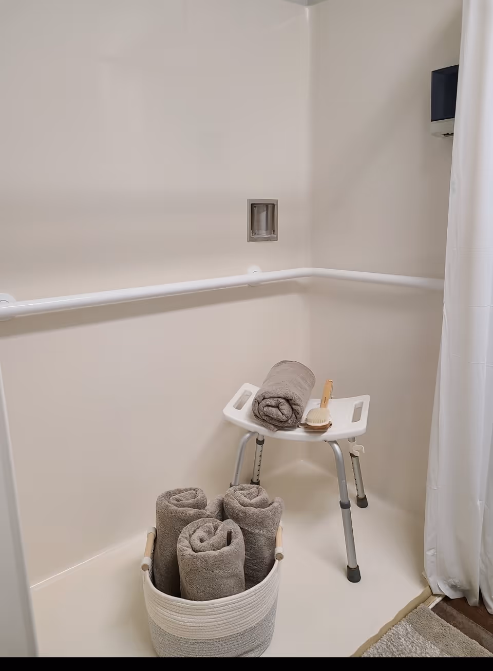 Accessible shower area with grab bars, a shower chair holding a rolled towel and brush, and a basket of rolled towels on the floor.