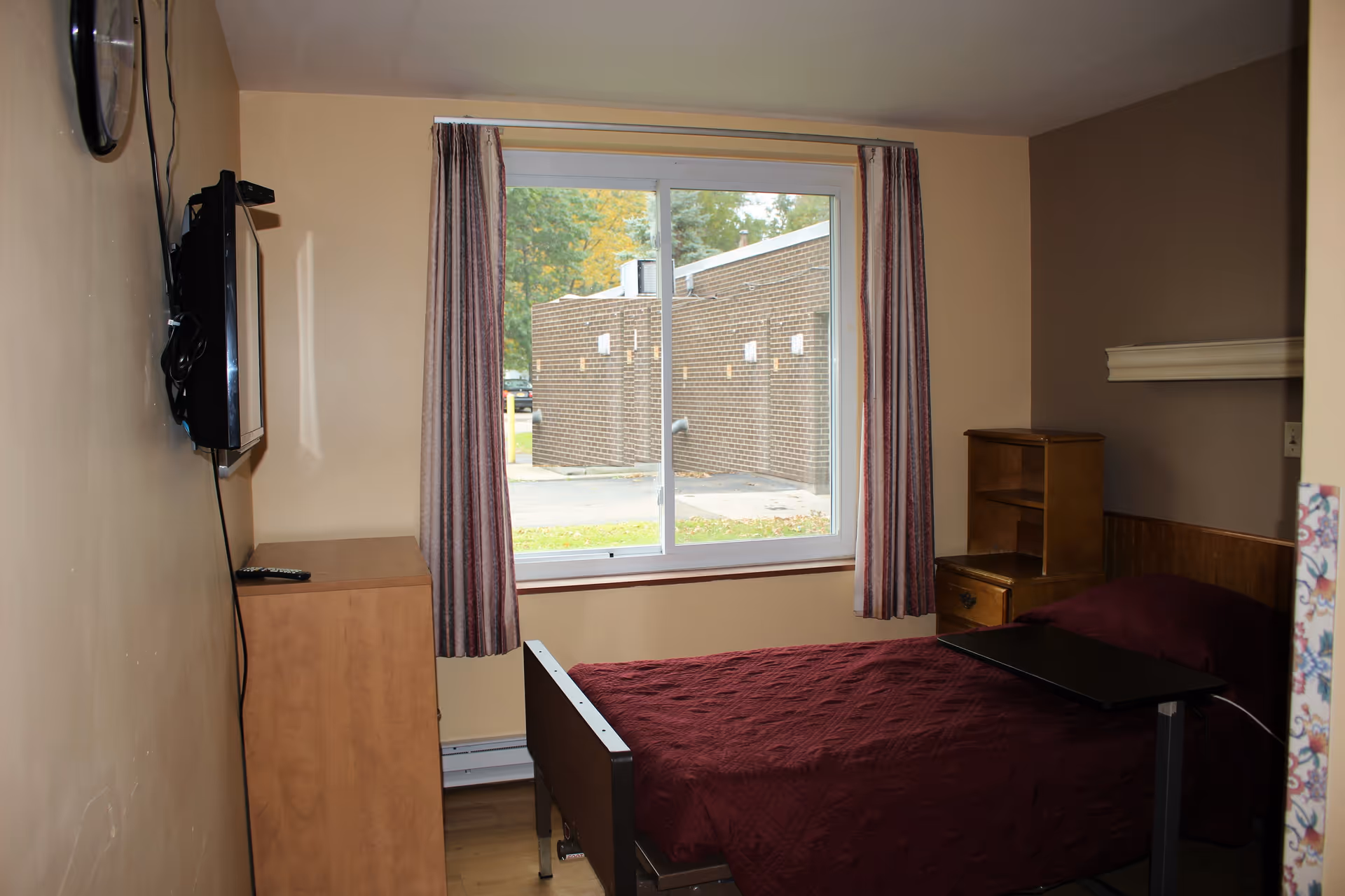 A small bedroom with a single bed covered in a dark red quilt. There is a wooden nightstand with shelves and a drawer next to the bed. A flat-screen TV is mounted on the wall opposite the bed, above a wooden dresser. A window with striped curtains shows an outside view of a brick building and some greenery.