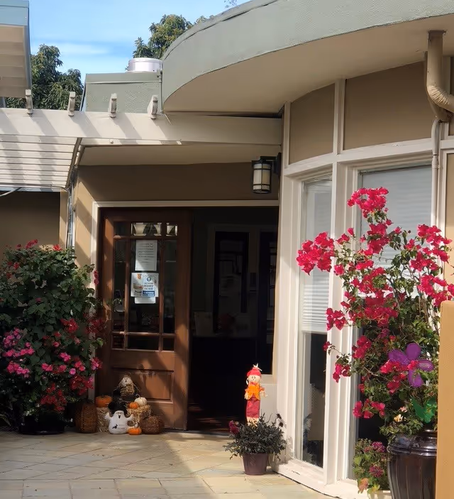 Front entrance of a building with a glass door, potted flowering plants, and autumn decorations.