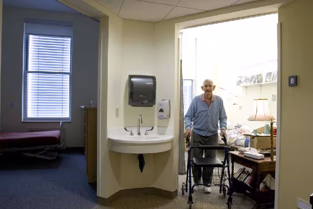 An elderly man using a walker stands in the doorway of a well-lit room with a bed, lamp, and personal items. To the left of the doorway is a wall-mounted sink with a paper towel dispenser and soap dispenser. Another bed is visible in a dimly lit adjacent room with a window covered by blinds.