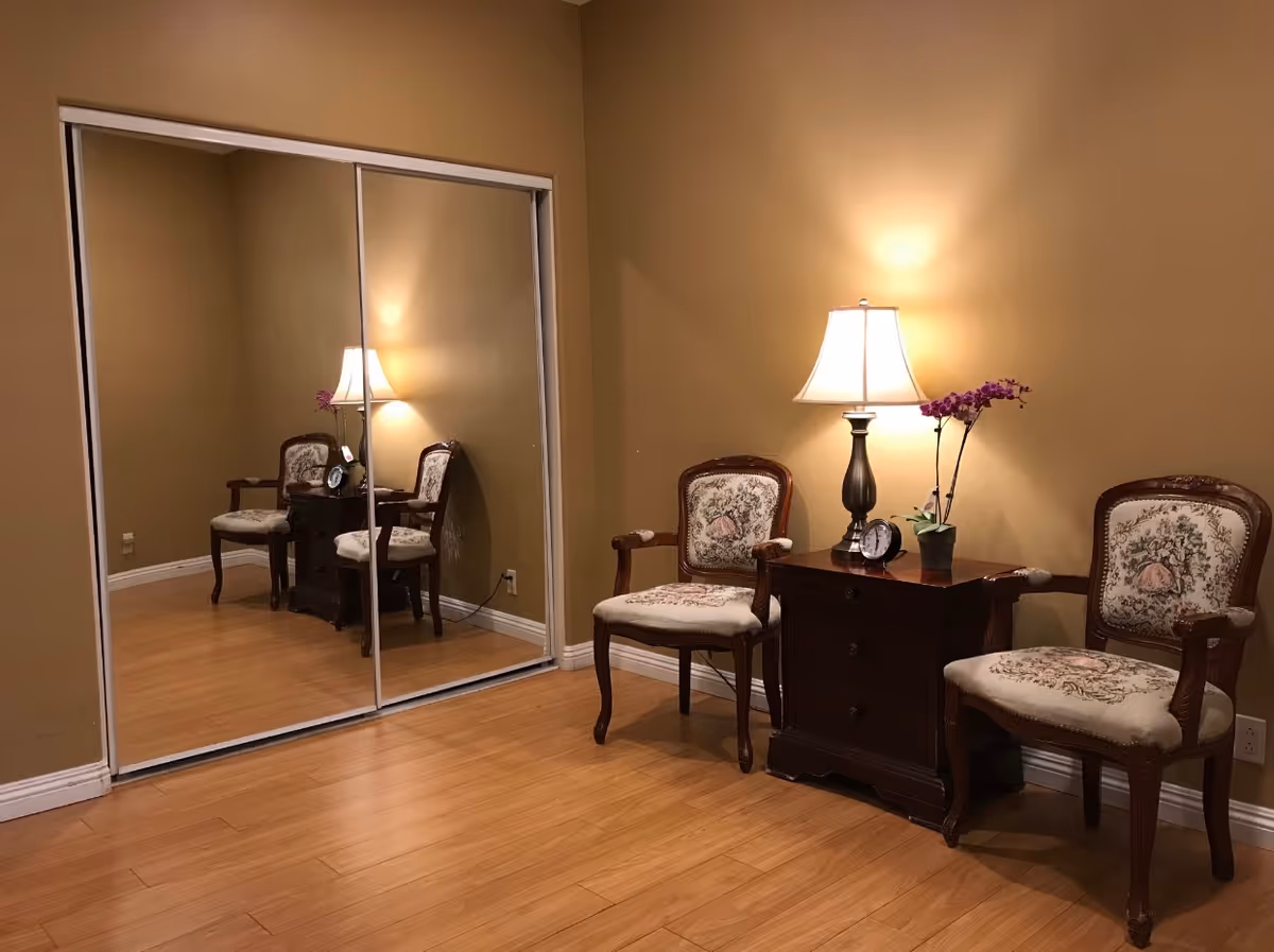 Two upholstered chairs and a wooden side table with a lamp and orchid in a small sitting area with mirrored closet doors and hardwood floor.
