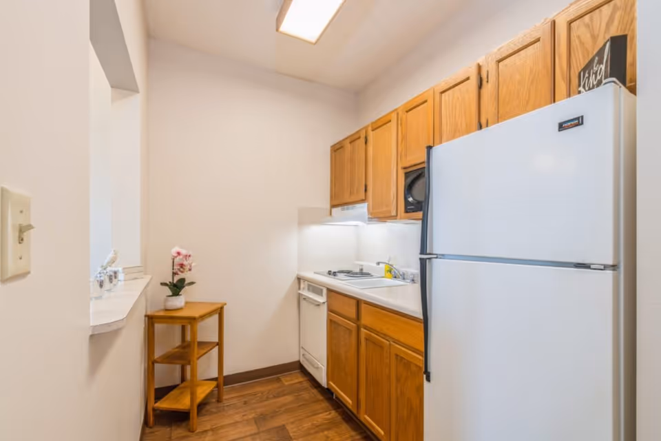 A small kitchen area with wooden cabinets, a white refrigerator, a white dishwasher, a sink, and a stove. There is a small wooden corner shelf with a potted orchid on it. The walls are light-colored, and the floor has a wood finish.