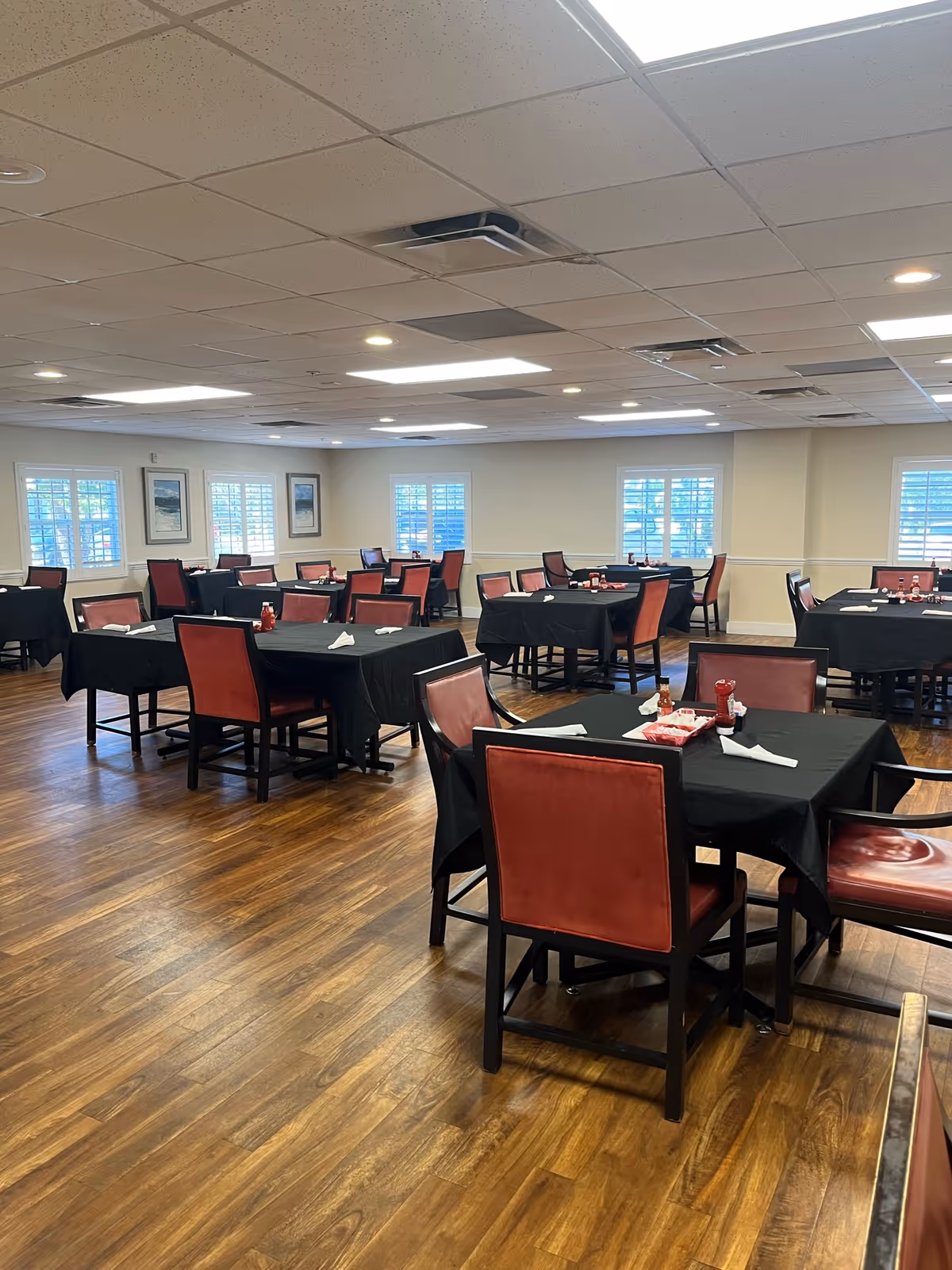 A dining room with multiple tables covered in black tablecloths and surrounded by red cushioned chairs. The room has wooden flooring, white walls with windows featuring white shutters, and ceiling lights providing bright illumination. Some tables have napkins and condiments placed on them.