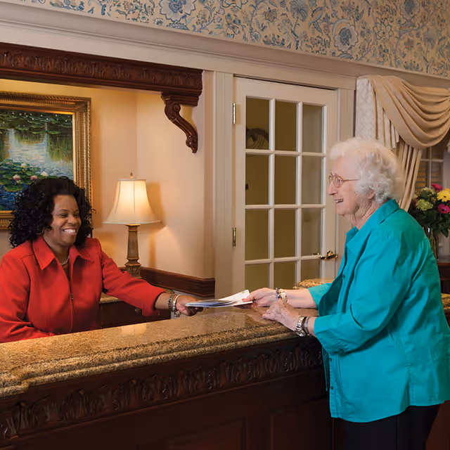 An elderly woman in a teal jacket is handing papers to a smiling receptionist in a red jacket across a polished wooden reception desk in a warmly decorated interior with floral wallpaper, a table lamp, and a framed painting.