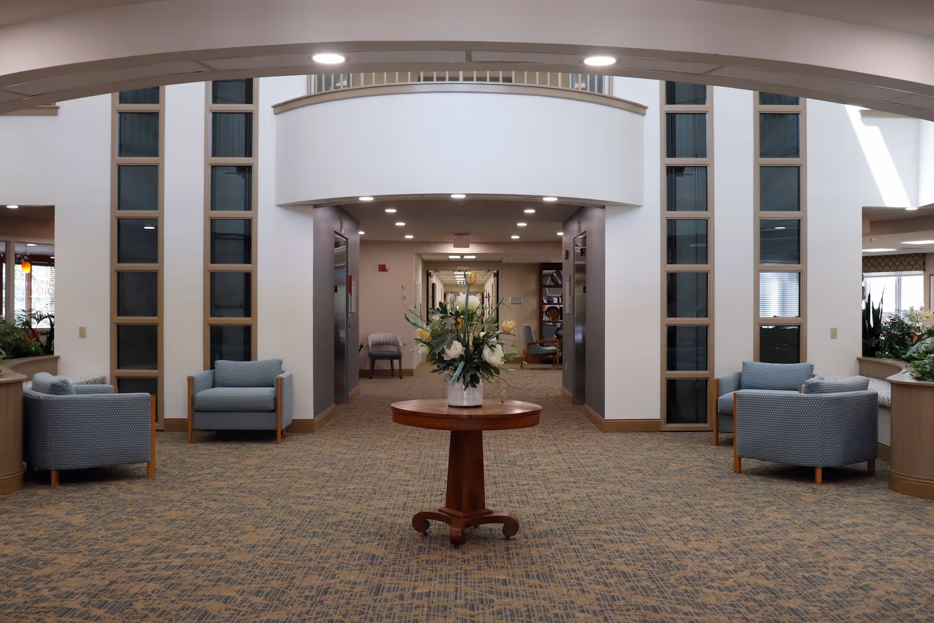 Spacious lobby atrium with a round table and floral arrangement at the center, upholstered chairs and elevators in the background.