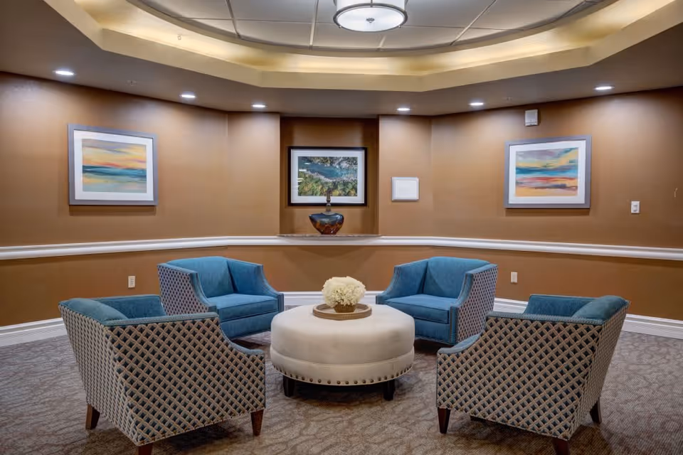 A cozy seating area with four blue upholstered armchairs arranged around a round white ottoman with a flower centerpiece, set against warm brown walls adorned with three framed landscape paintings and soft overhead lighting.