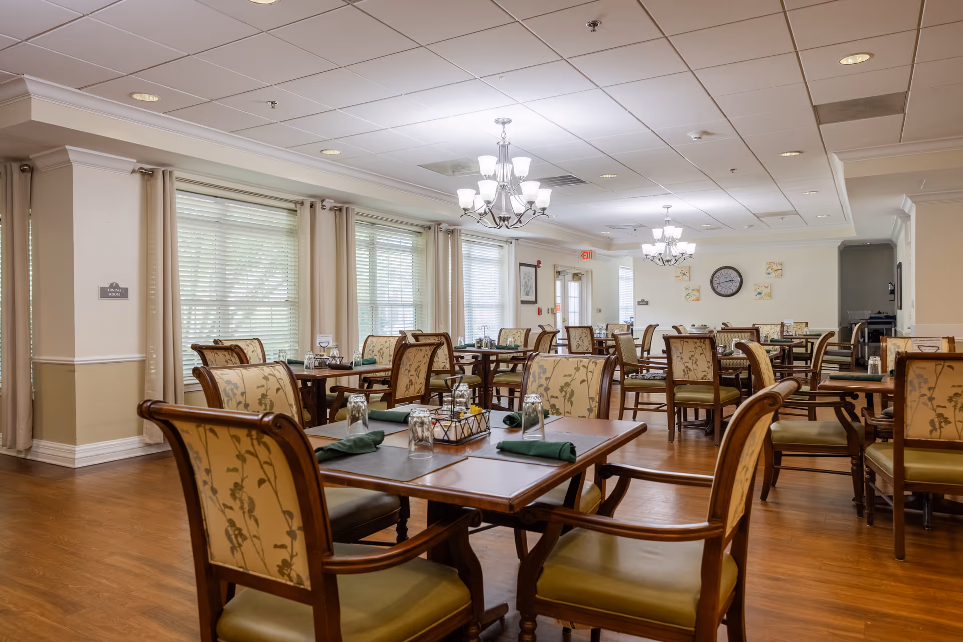 A spacious dining room with multiple wooden tables and cushioned chairs featuring floral upholstery. The tables are set with green napkins and upside-down glasses. Large windows with beige curtains allow natural light to fill the room. Elegant chandeliers hang from the ceiling, and a clock and framed pictures are visible on the far wall.