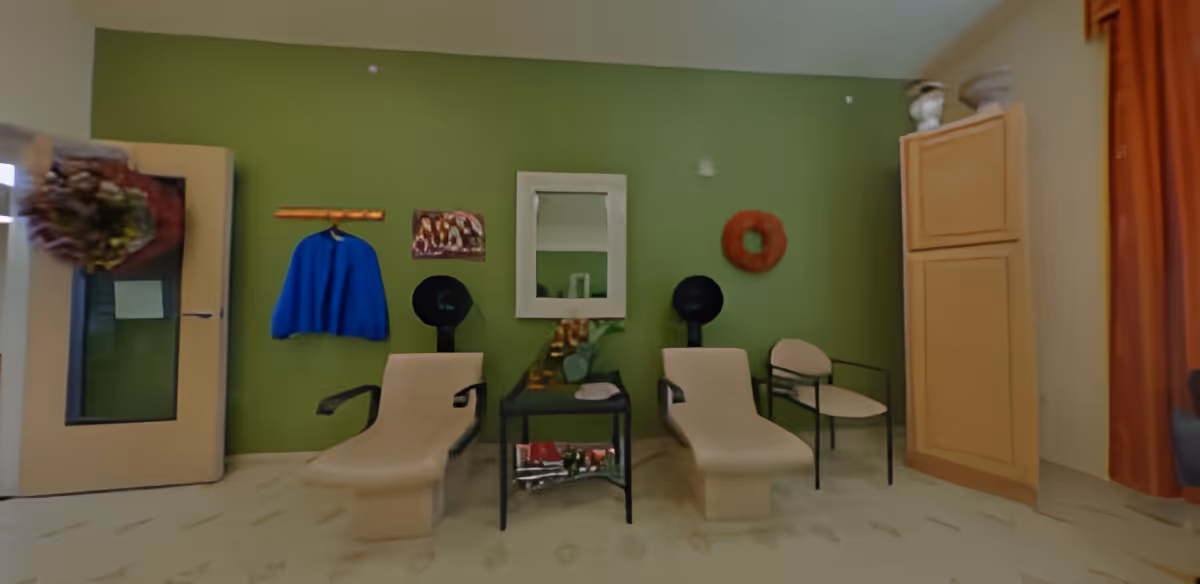 Small salon-style room with two reclining styling chairs and hooded hair dryers against a green wall with a mirror and side table between them.