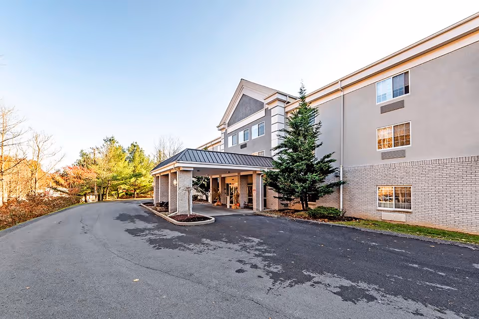 Exterior view of a senior living facility building with a covered entrance, surrounded by trees and a paved driveway under a clear sky.