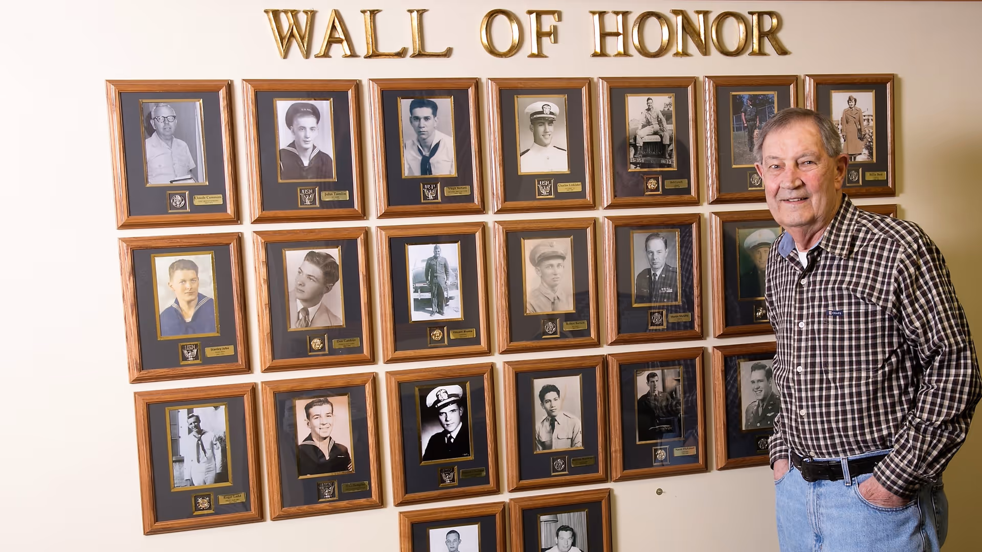 An elderly man stands next to a 'Wall of Honor' displaying framed black-and-white and color photographs of individuals, each with a small plaque beneath. The wall is painted a light color and the words 'WALL OF HONOR' are displayed in large gold letters above the photos.