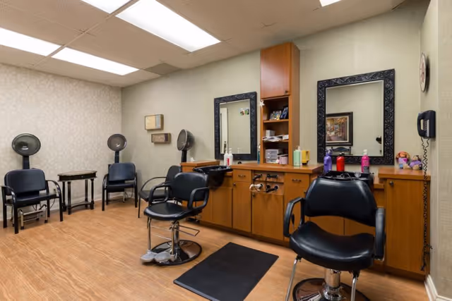 Interior view of a hair salon area in a senior living facility with two black salon chairs in front of a wooden counter with mirrors, hair care products, and hair dryers mounted on the wall. The room has beige walls and wood flooring.