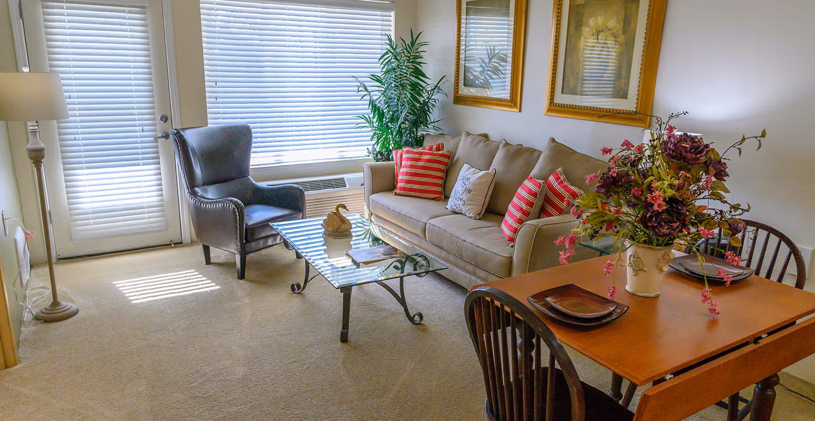 Bright living room with a beige sofa and striped pillows, glass coffee table, armchair, small dining table with a floral centerpiece, and blinds-covered door and window.