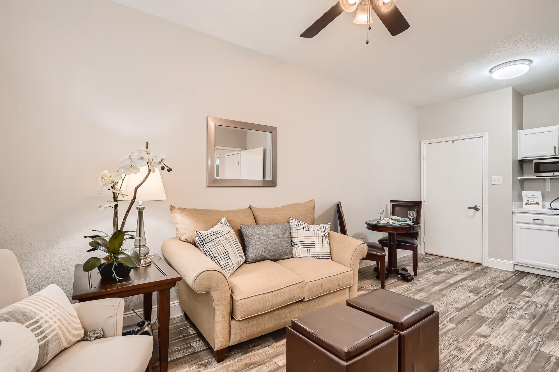 A cozy living room area with a beige loveseat adorned with three decorative pillows, a wooden side table with a lamp and a white orchid plant, a beige armchair with a patterned pillow, two brown leather ottomans, a small round dining table set for two with wine glasses and plates, and a partial view of a white kitchen with cabinets and a microwave. The room has wood-patterned flooring, a ceiling fan with lights, and a wall mirror above the loveseat.