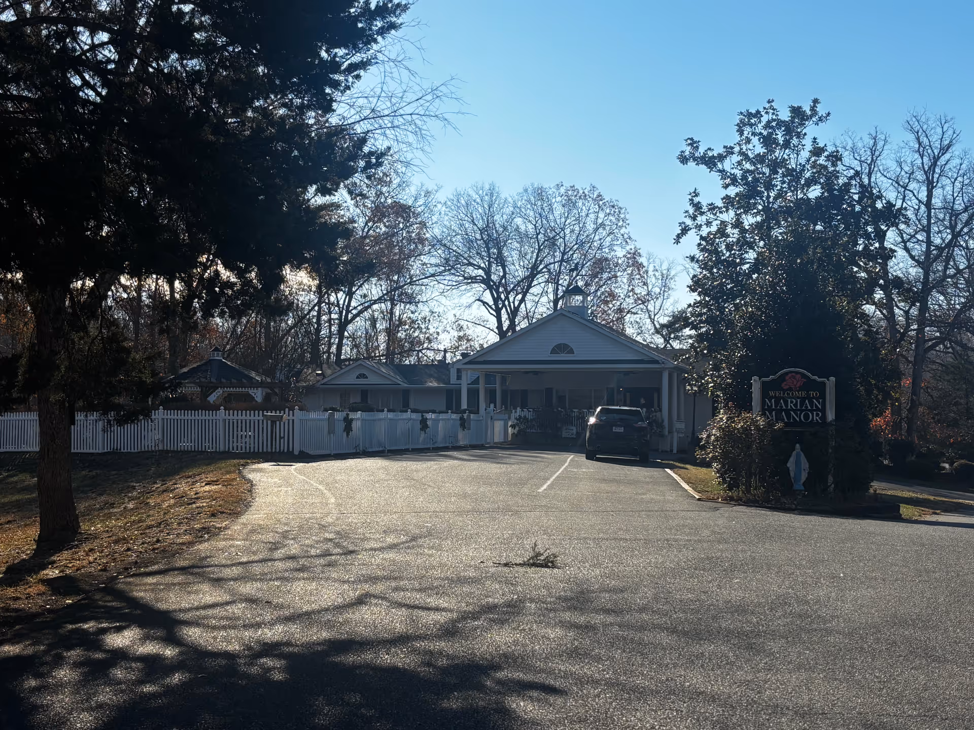 Front entrance of Marian Manor with a driveway, parked car, white picket fence and a sign.
