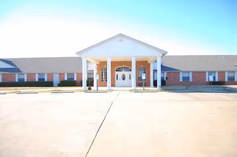 Single-story brick senior living facility with a white portico and expansive front parking.
